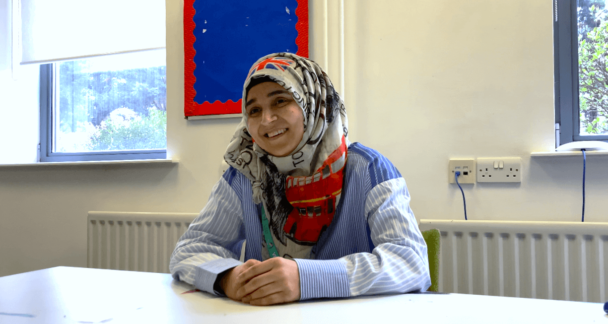A person wearing a patterned headscarf and striped shirt sits at a table, smiling, in a room with white walls and a blue bulletin board, where an Employability Programme session is taking place. | SEN Schools