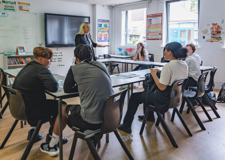 A teacher stands at the front of a classroom, engaging with six seated students around a table. | SEN Schools