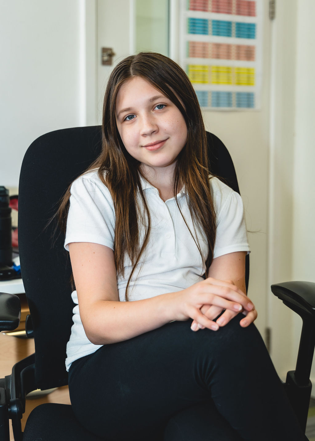 A young girl with long brown hair sits in a swivel chair, wearing a white shirt and black trousers, looking at the camera. | SEN Schools