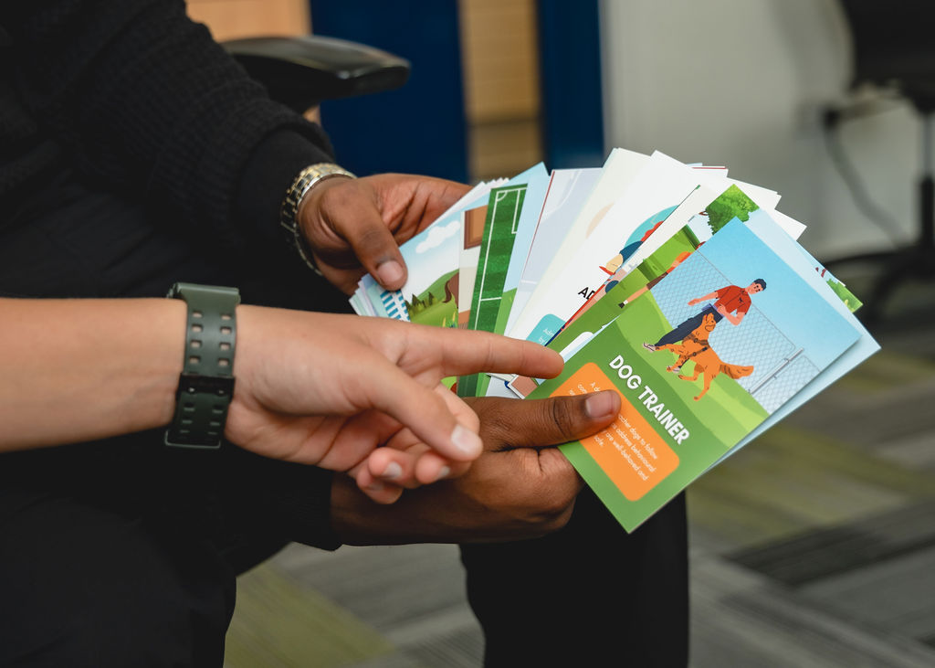 Two people are seated and holding a collection of colorfully illustrated cards, with one card labeled "Dog Trainer. | SEN Schools
